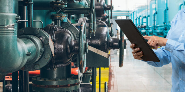 Person with a tablet inspecting a row of industrial pipelines and valves in a technical facility.